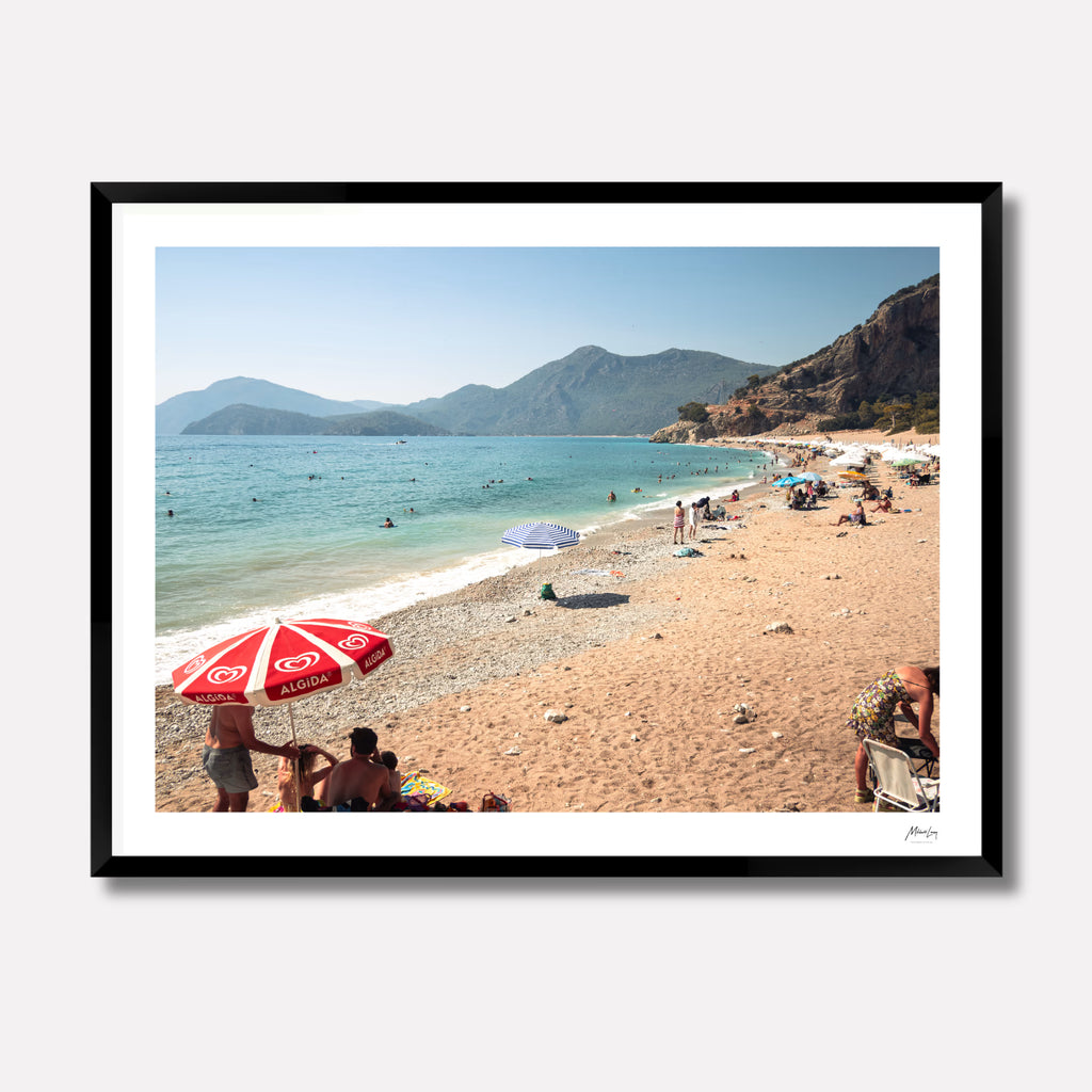 Fine art photography titled 'Magnums' by Venturer Studios, featuring a sunlit beach in Western Turkey dotted with vibrant, colorful umbrellas. The bright coastal scene is captured in a deep black gallery frame.
