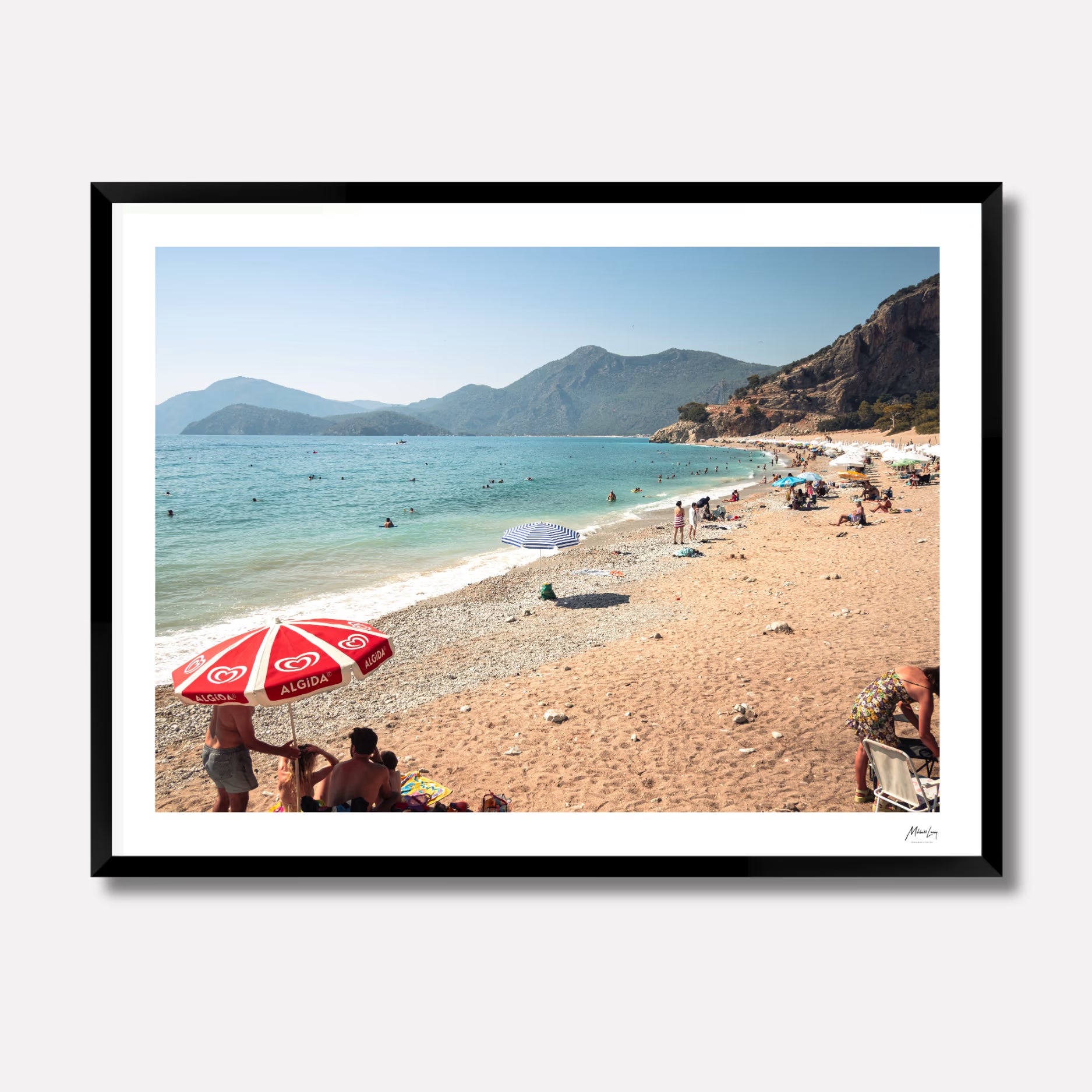 Fine art photography titled 'Magnums' by Venturer Studios, featuring a sunlit beach in Western Turkey dotted with vibrant, colorful umbrellas. The bright coastal scene is captured in a deep black gallery frame.
