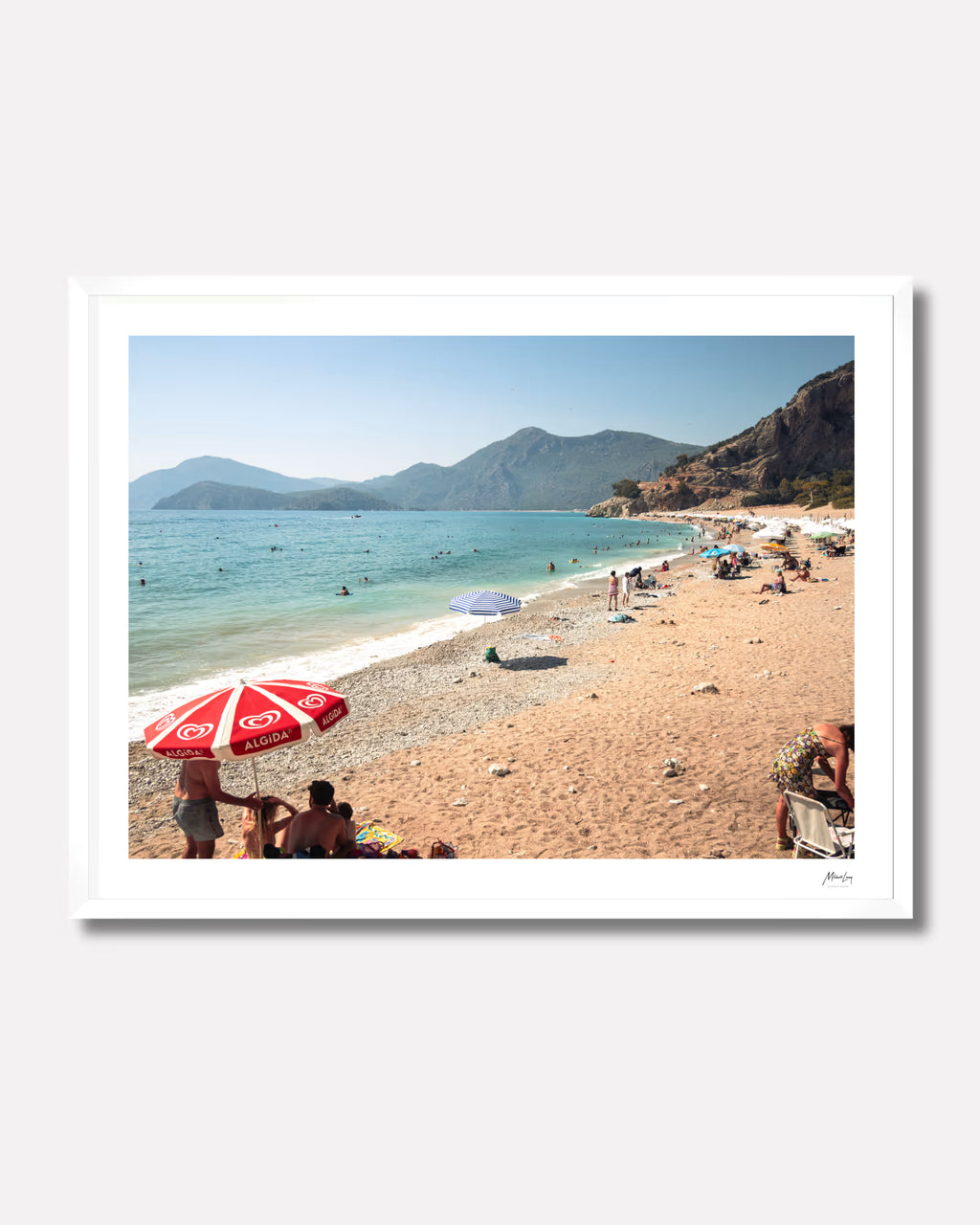 White framed photograph of a beach scene with people, umbrellas, and mountains.