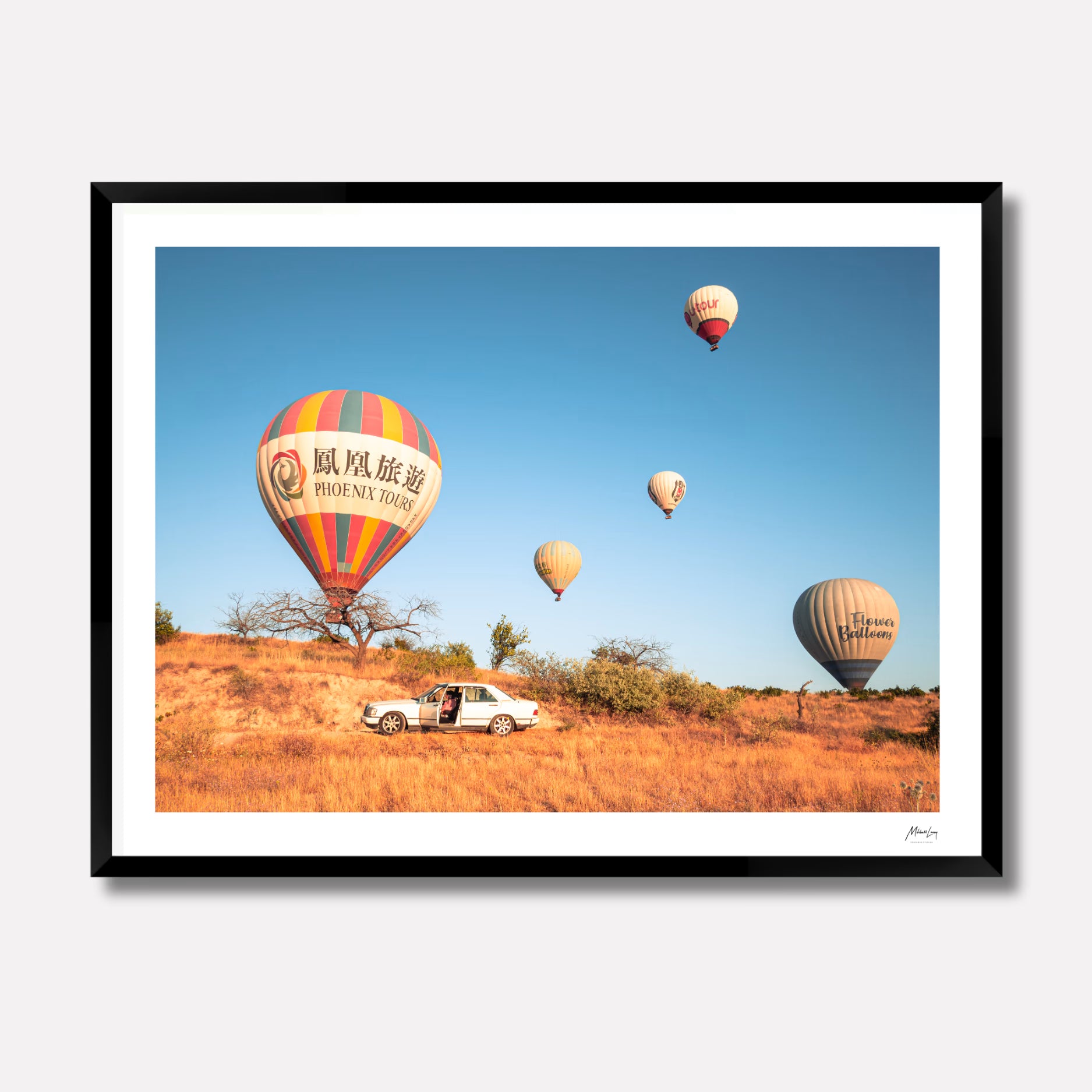 Fine art photography titled 'Parked,' featuring colorful hot air balloons floating over the rocky desert landscape of Cappadocia, Turkey, with a vintage car parked in the foreground. The print is displayed in a 4cm deep matte black gallery frame.