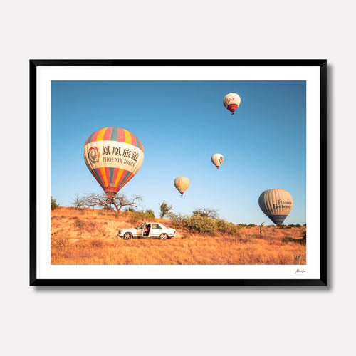 Fine art photography titled 'Parked,' featuring colorful hot air balloons floating over the rocky desert landscape of Cappadocia, Turkey, with a vintage car parked in the foreground. The print is displayed in a 4cm deep matte black gallery frame.
