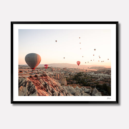 Fine art photography titled 'Rising' by Venturer Studios, capturing multiple hot air balloons ascending over the Cappadocia landscape during a golden sunrise. The print is showcased in a deep black gallery frame.